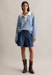 Camicia a maniche lunghe a righe blu e bianche con colletto e tre bottoni, abbinata a una gonna plissettata in denim scuro e stivali con tacco marroni.