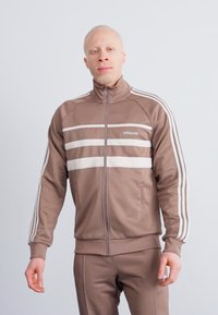 Brown athletic jacket with white stripes, zip front, high collar, and smooth texture. Worn with matching pants, showcasing minimalist design.