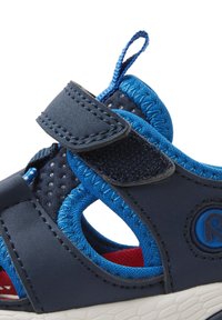 Sinine ja tumesinine sandaal, millel on reguleeritav Velcro rihm, tekstureeritud pinnad ja perforeeritud küljed. Valge kummist tald punase sisetallaga.