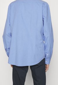 Camisa de manga larga azul claro hecha de algodón. Presenta un dobladillo redondeado, puños con botones y una textura suave. La vista trasera no muestra logotipos visibles.
