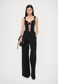Fekete hálós bodysuit formázott pántokkal és széles fekete nadrággal, amely zsebekkel rendelkezik, kiegészítve egy kis fekete kézi táskával és magassarkú cipővel.
