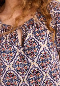 Kukallinen blusa, jossa on sinisiä, punaisia ja kermaisia värejä, kauluksessa solmimisdetalji ja joustavat röyhelöreunat.