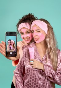 Två personer med ansiktsmasker poserar för en selfie. En av dem har på sig en rosa sidenblus med prickar och håller i en rosa tube märkt "Handy."