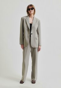 Lys grå skreddersydd blazer med knepping i nakken, enkel knapp luke, og frontlommer, kombinert med matchende bukser og svart topp.