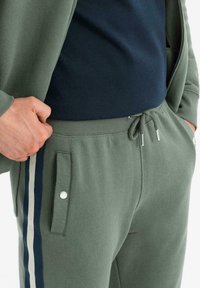 Homme portant un pantalon de survêtement vert olive avec des bandes latérales bleu marine et blanches, un t-shirt noir, et une veste assortie vert olive.