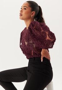 Lila genomskinlig blus dekorerad med blommiga applikationer, med puffärmar och avslappnad passform, ihop med svarta åtsittande byxor.