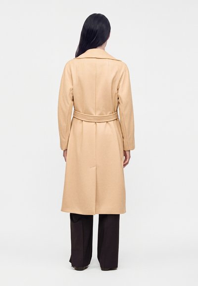 Femme aux longs cheveux foncés, portant un long manteau beige ceinturé et un pantalon noir à jambes larges, debout dos au spectateur sur fond blanc.