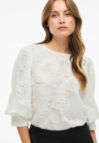 Blouse blanche texturée avec un motif tissé, col rond et manches bouffantes trois quarts, associée à un pantalon noir.
