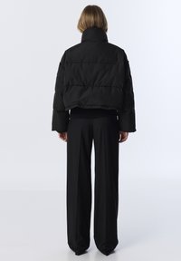 Veste en duvet noire courte avec un col montant et une texture matelassée, associée à un pantalon noir ample et long. Fond uni.