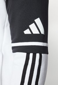 Schwarz-weißer Sweatshirt mit drei vertikalen schwarzen Streifen, einem weißen Adidas-Logo auf dem Ärmel und gerippten Handgelenkbündchen.