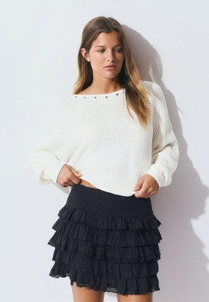 Suéter blanco de punto con corte holgado y textura acanalada; cuenta con botones decorativos de metal. Falda negra de volantes en capas con una cintura fruncida.