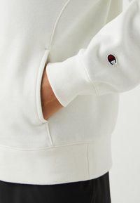 Sudadera blanca con puños y dobladillo acanalados, que presenta un bolsillo lateral y un pequeño logo en la manga. Tela suave y texturizada.