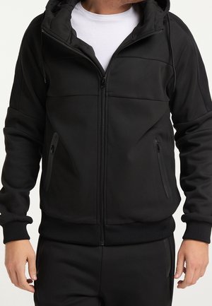 Homme portant un sweat à capuche noir zippé avec des poches zippées et un t-shirt blanc en dessous, debout les bras détendus.