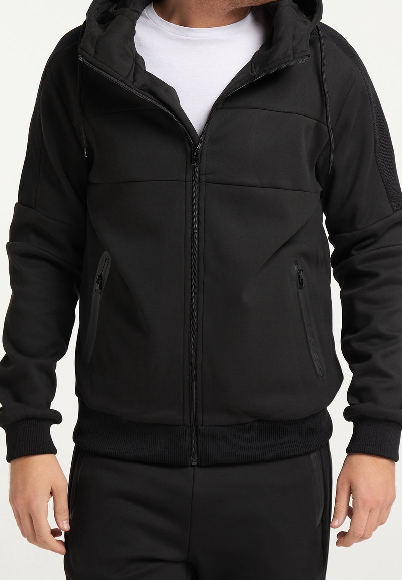 Homme portant un sweat à capuche noir zippé avec des poches zippées et un t-shirt blanc en dessous, debout les bras détendus.