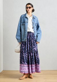 Veste en jean portée sur une chemise blanche, associée à une jupe maxi violette à motifs avec des accents roses, et des bottines brunes. Petit sac à main blanc.