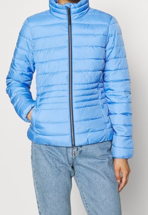 Personne portant une veste matelassée bleu clair avec une fermeture éclair devant et les mains dans les poches latérales, associée à un jean bleu clair.