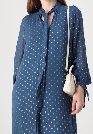 Vrouw in een blauwe lange-mouwen jurk met zilveren stippen en een detail met een necktie, die een kleine witte handtas op haar schouder draagt.
