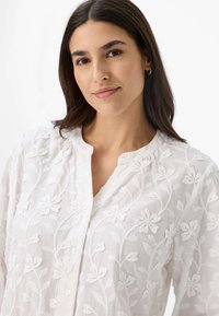 Weiße, bestickte Bluse mit floralen Mustern, rundem Halsausschnitt und Knopfschluss. Weiche Textur, leichtes Material und lässige Passform.