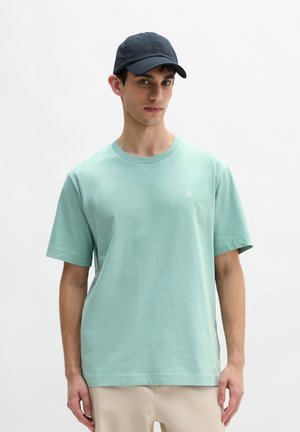 Młody mężczyzna w jasnozielonej koszulce i granatowej czapce, stojący na tle jednolitego białego tła.