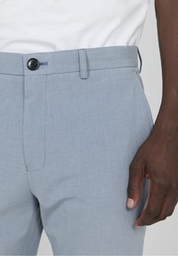 Světle modré kalhoty z texturované tkaniny s plochým předním dílem, vybavené černým knoflíkem a bočními kapsami. Detailní záběr na pás a ruku.