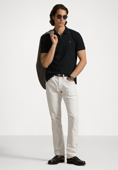 Svart polo skjorte med korte ermer, hvite jeans og svarte loafers. Holder en lys grå jakke. Bruker runde solbriller og et armbåndsur.