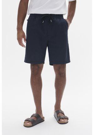 Homme portant un short bleu marine à cordon, un t-shirt blanc et des sandales noires à boucle, debout les mains dans les poches sur un fond blanc.