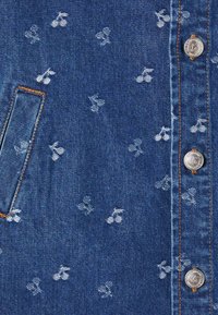 Tessuto di denim blu con un motivo a ciliegie sparse in bianco. Include una tasca e un dettaglio con bottone argentato e cuciture arancioni.