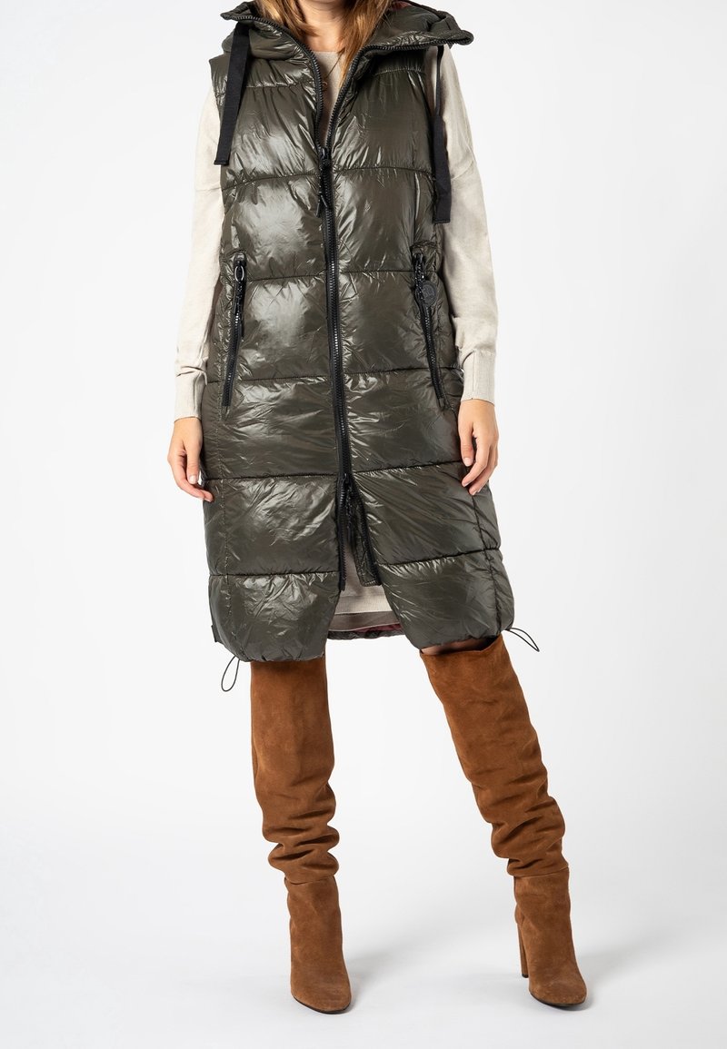 Femme portant une veste matelassée sans manches vert olive longueur genou avec fermeture éclair devant et bottes hautes en daim marron.