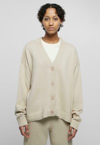 Beige oversized cardigan with a V-neck, large buttons, and ribbed texture. Features dropped shoulders and wide sleeves, layered over a light top.