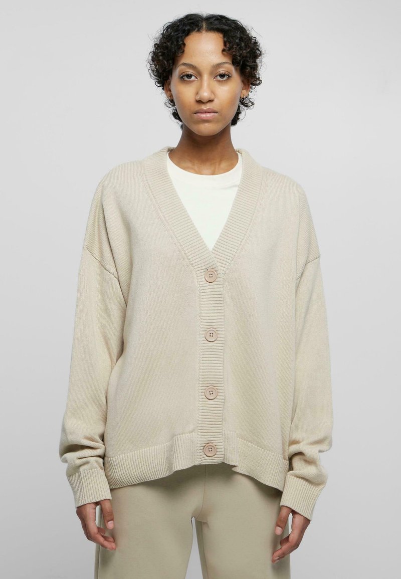 Beige oversized cardigan with a V-neck, large buttons, and ribbed texture. Features dropped shoulders and wide sleeves, layered over a light top.