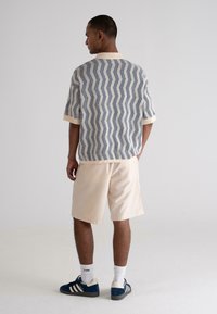 Cămașă cu mânecă scurtă pentru bărbați, albă, cu un model în zigzag albastru, asortată cu pantaloni scurți de culoare bej deschis. Poartă adidași albaștri cu accente albe.
