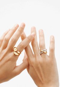 Dos manos con anillos: uno de forma alargada de oro con tres acentos redondeados y otro de banda entrelazada de plata y oro. Texturas suaves, diseño minimalista.