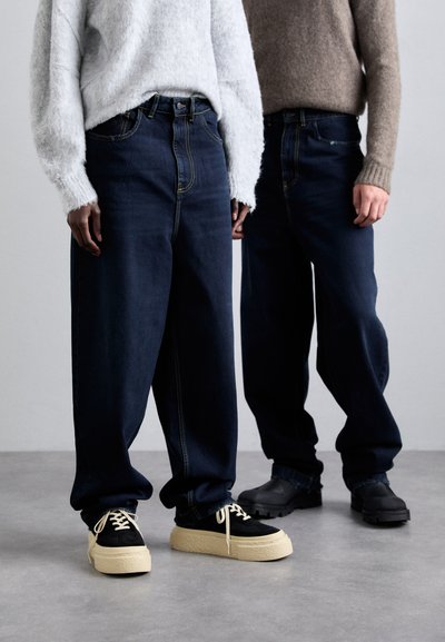 Deux personnes portant des jeans en denim foncé amples avec des coutures visibles. L'une porte un pull en laine moelleux gris clair, l'autre un pull en tricot marron.