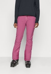 Roze geïsoleerde skibroek met een recht model, zijzakken en versterkte onderbenen, gecombineerd met zwarte skischoenen.