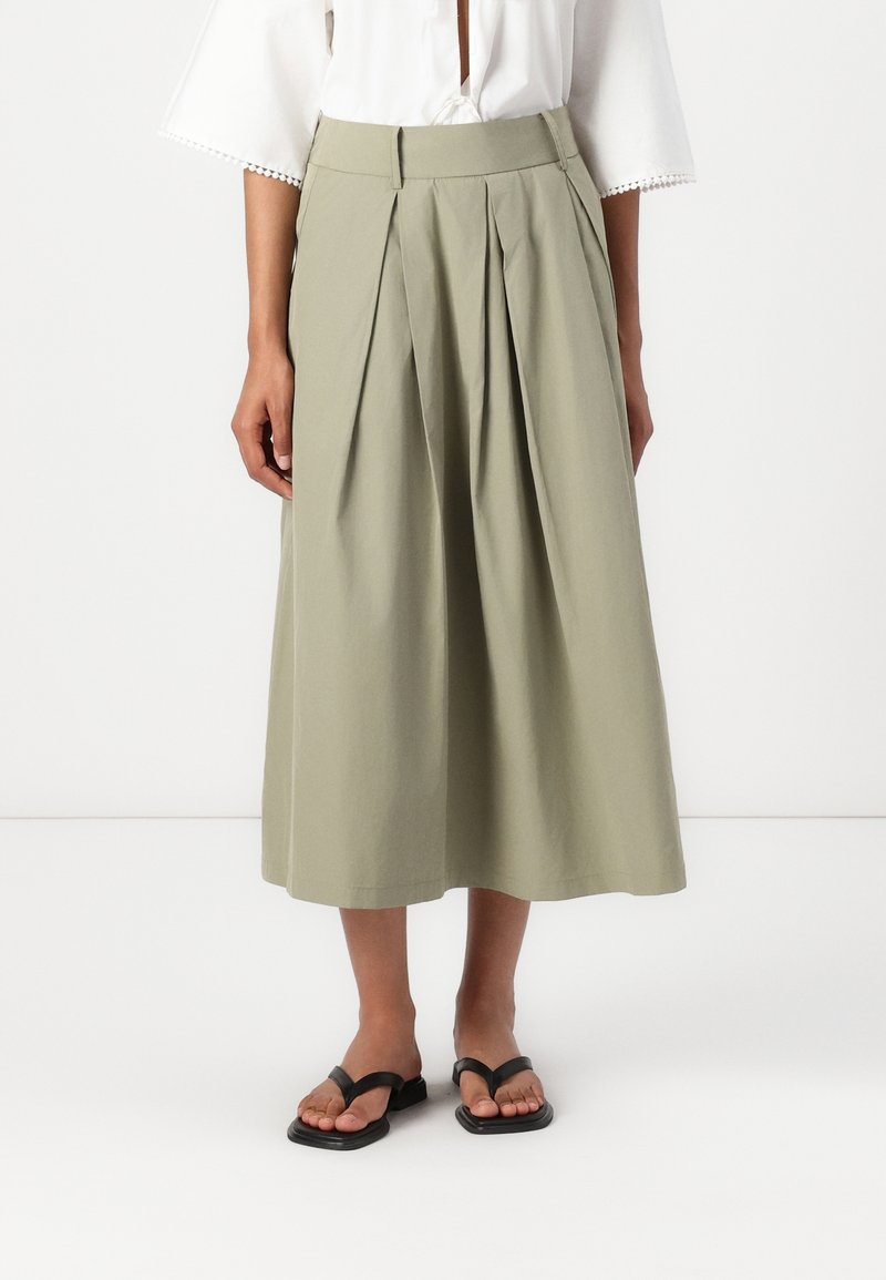 Femme portant une jupe midi plissée vert olive clair, un chemisier blanc à manches trois-quarts et des sandales noires à bout carré.