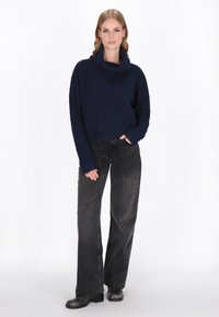 Vrouw staat met hand in de zak, gekleed in een marineblauwe coltrui, wijde zwarte jeans en zwarte laarzen, op een witte achtergrond.