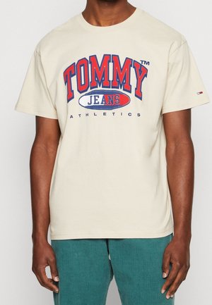 Homme portant un T-shirt beige à manches courtes avec un logo "Tommy Jeans Athletics" rouge et bleu et un pantalon texturé bleu sarcelle.