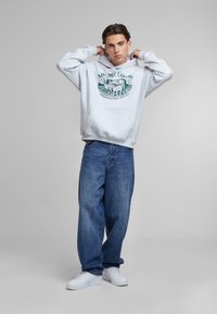 Gray hoodie with a green graphic print, front pocket, and ribbed cuffs, paired with loose-fitting blue jeans and white sneakers.