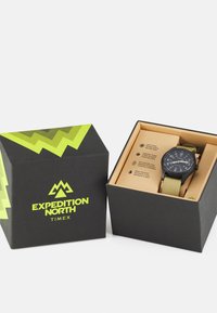 Schwarze Uhr mit grünen Akzenten und einem Stoffarmband, präsentiert in einer schwarzen Box mit grünem Zickzackmuster und umweltfreundlichen Details.