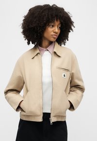 Γυναίκα με σγουρά μαλλιά, φοράει μπεζ μπουφάν με φερμουάρ πάνω από λευκή μπλούζα και ροζ πουκάμισο με γιακά, με τα χέρια στις τσέπες, κοιτάζει προς το πλάι.