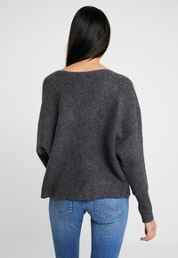 Pull en tricot gris à coupe décontractée et manches longues, avec un col large et des poignets côtelés, associé à un jean bleu.