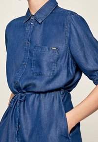 Jeansskjolklänning i mörkblått med krage, korta ärmar, en bröstficka och dragsko i midjan för justerbar passform.