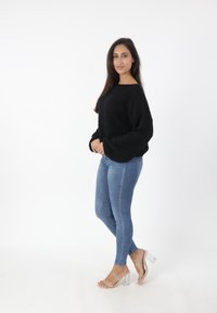 Zwarte, gestructureerde trui met brede mouwen, in combinatie met strakke blauwe jeans en doorzichtige sandaaltjes met hak. Model staat tegen een witte achtergrond.