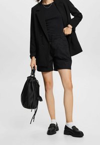 Czarna marynarka z podwójnymi guzikami, czarny t-shirt, dopasowane czarne szorty, trzyma czarną torbę, w połączeniu z czarnymi mokasynami i białymi skarpetkami.