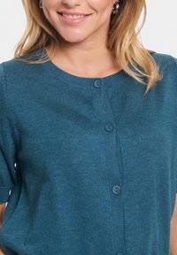 Mėlynas mezginys kardiganas su apvaliu iškirpimu ir trumpomis rankovėmis. Yra mygtukų uždarymas priekyje su atitinkančiais mygtukais ir braižyta tekstūra.