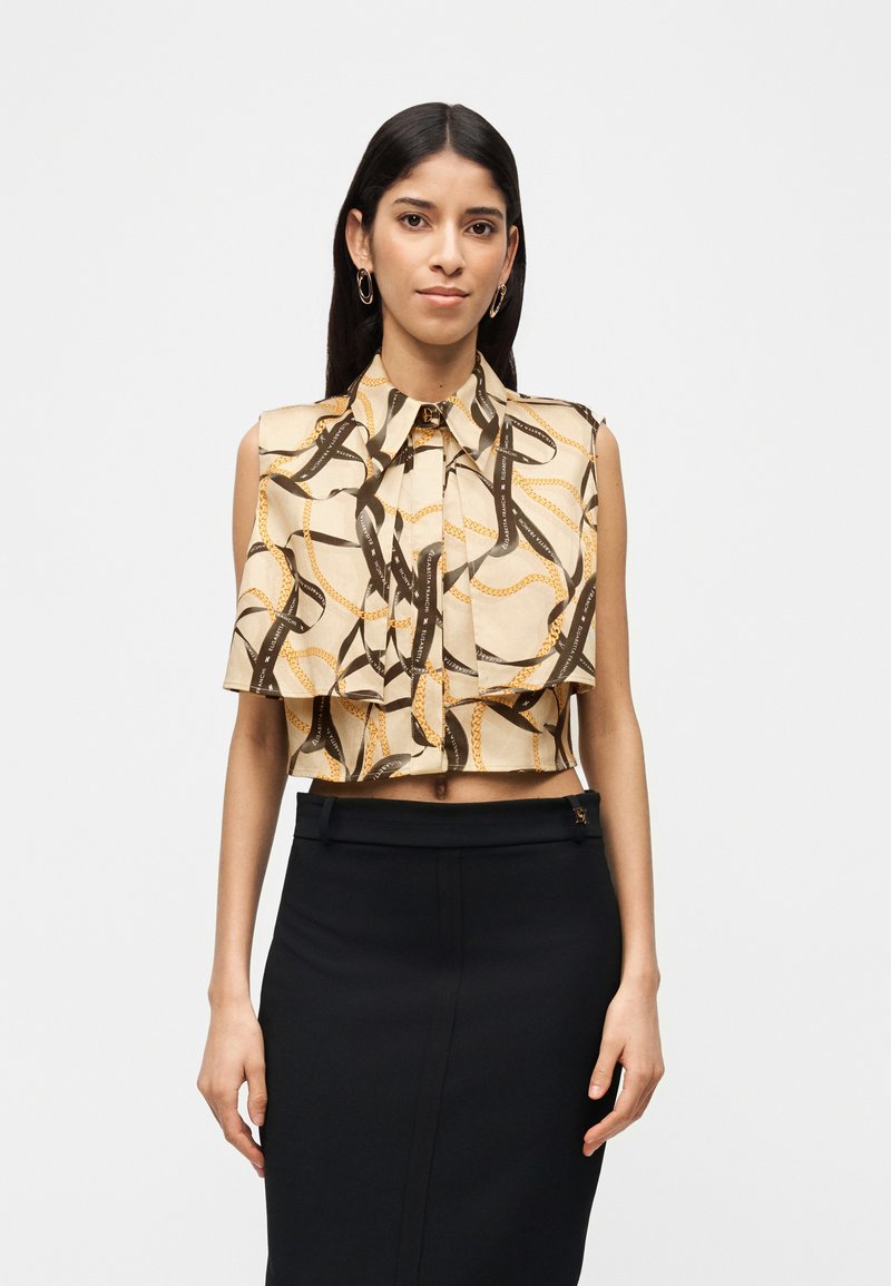 Γυναίκα με μπεζ αμάνικο μπλουζάκι με σχέδια, κομμένο στο ύψος του κορμού, και μαύρη ψηλόμεση φούστα, στέκεται μπροστά σε απλό λευκό φόντο.