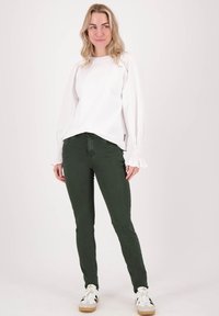 Witte lange mouwen top met gatherings aan de manchetten, gecombineerd met donkergroene skinny jeans. Model draagt witte sneakers met zwarte strepen.