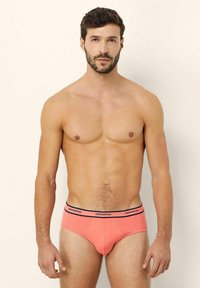 Vuxen man med kort mörkt hår och skägg som bär korallfärgade kalsonger med svart midjeband märkt "yamamay," stående mot en neutral bakgrund.