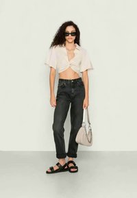 Camisa bege cortada com botões e mangas curtas, jeans pretos de cintura alta, sandálias pretas e uma mala cinza-claro com alça de corrente.