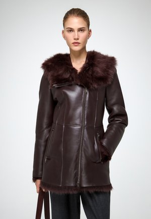 Veste en cuir marron avec un col en fourrure. Caractéristiques : fermetures éclair, poches latérales et coupe ajustée. Texture douce et design épuré.